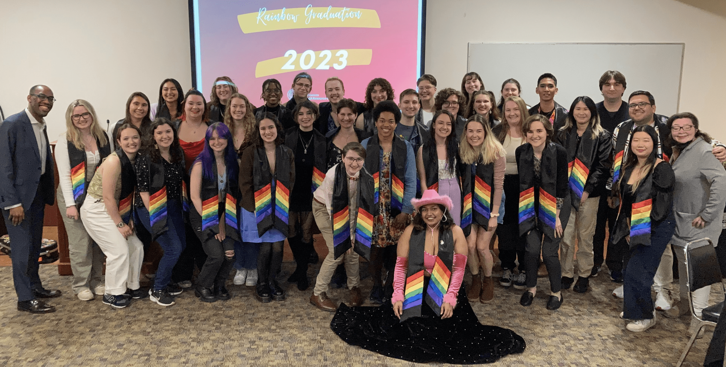 an image shows roughly 35 graduating seniors wearing their rainbow stoles in front of a screen Reading Rainbow graduation 2023.
